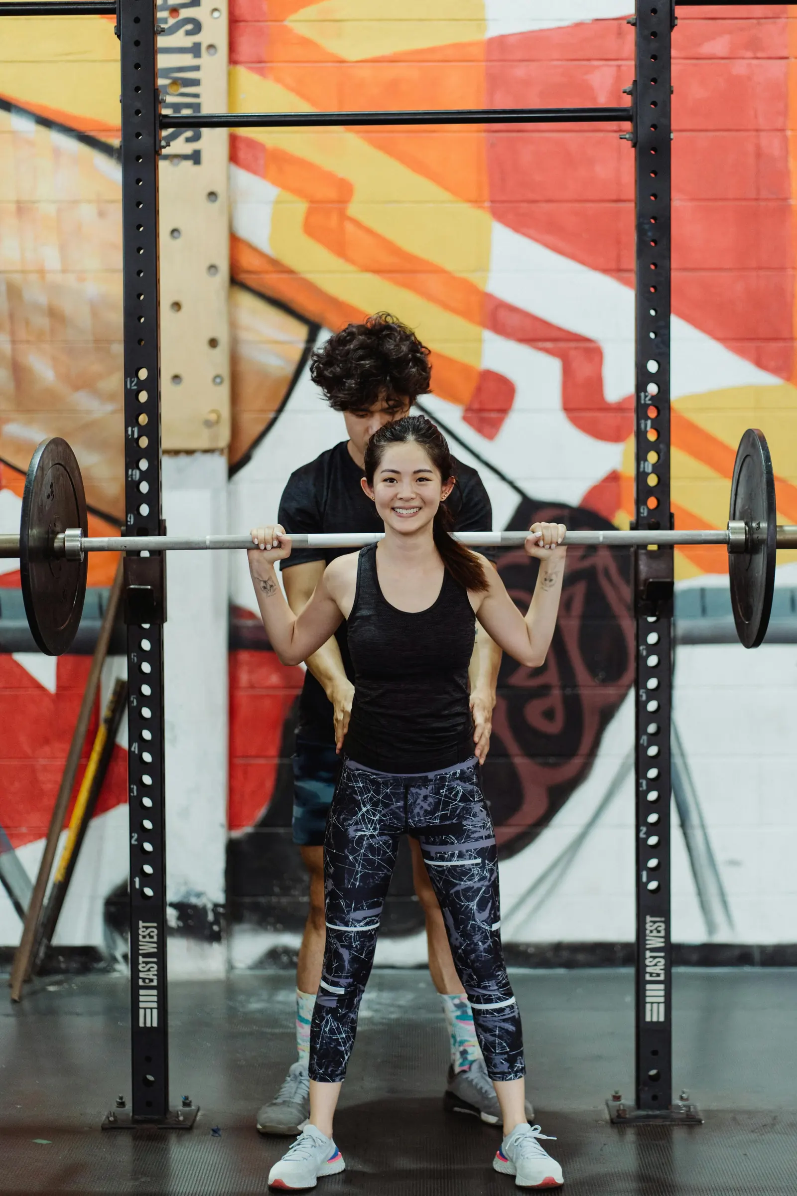 A woman squatting with a barbell in a rack in front of a painted wall while a trainer watches