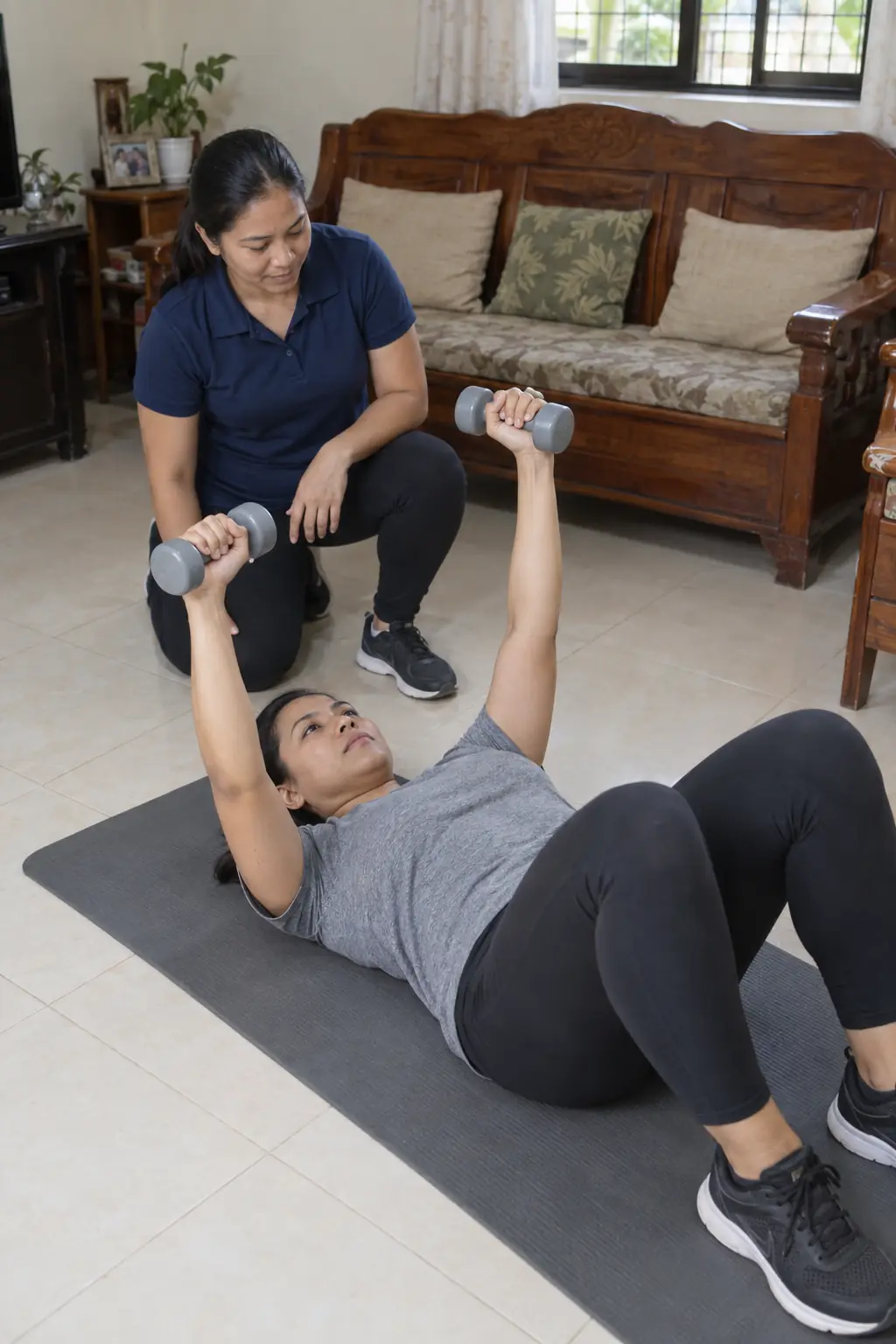 A Filipina woman lying on a mat pressing two dumbbells up from the floor, a Filipina coach kneeling at her head