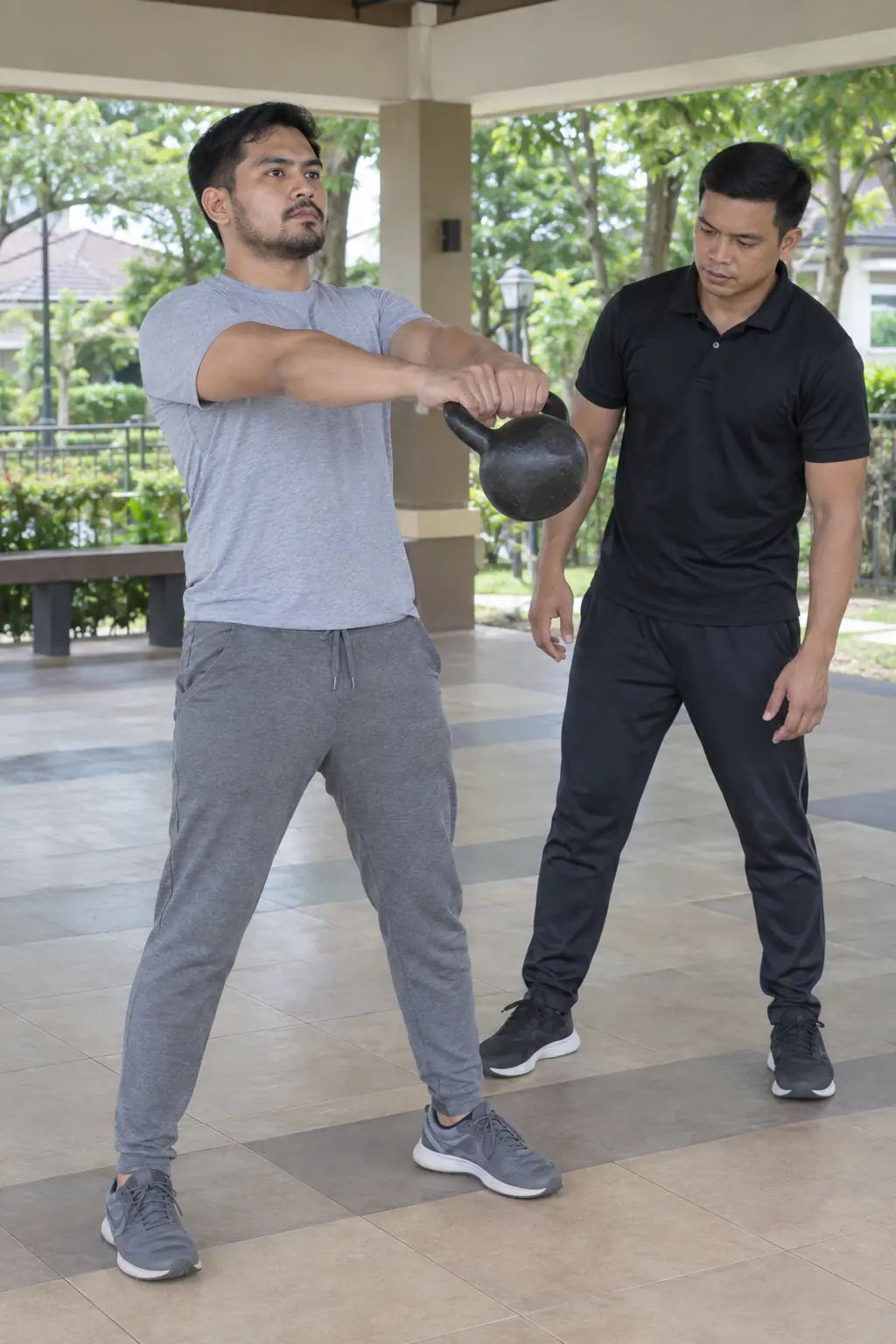 A Filipino man swinging a kettlebell to chest height in a covered clubhouse area while a Filipino coach watches