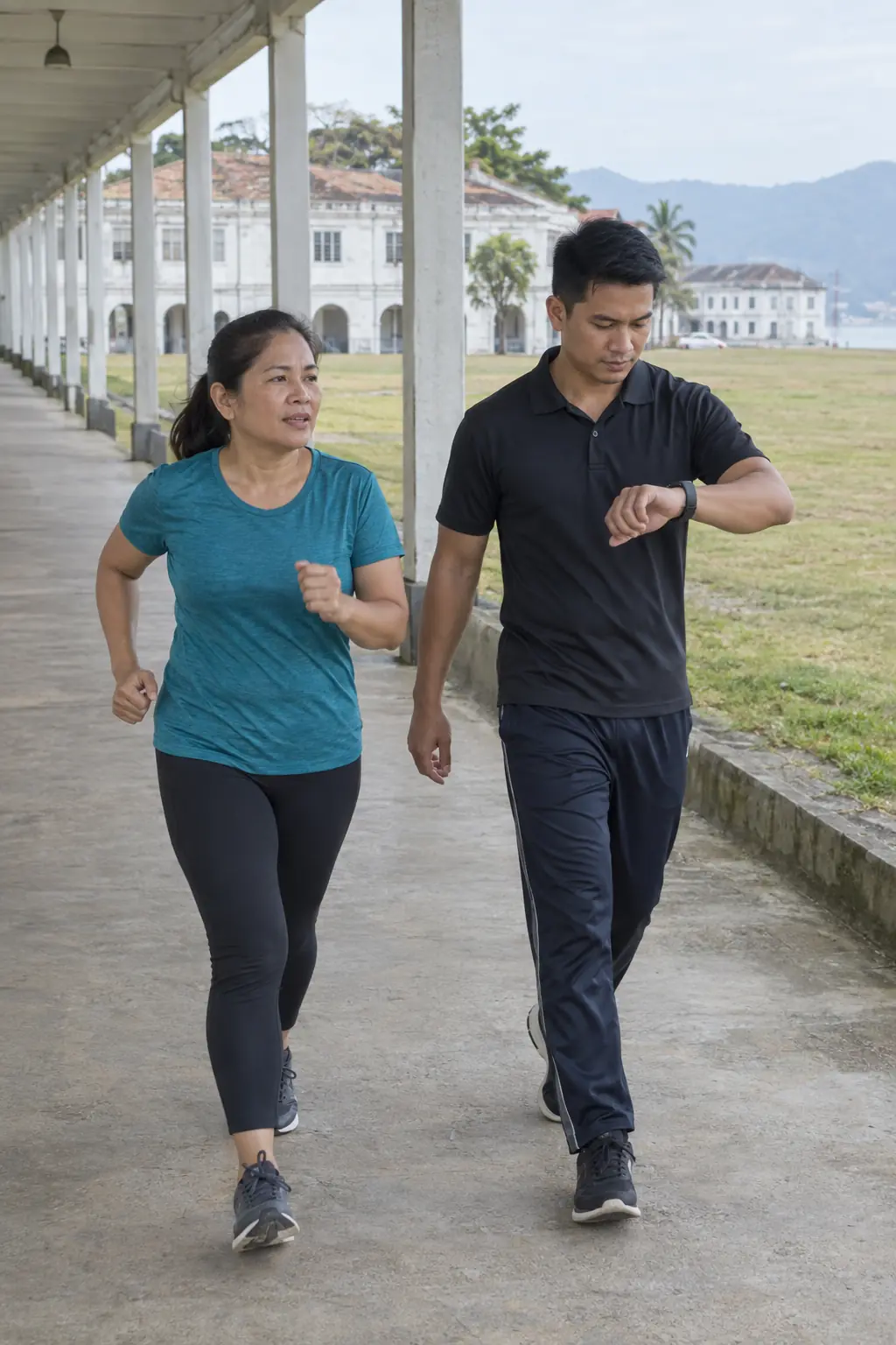 A Filipina woman power walking along a covered walkway beside a field, a Filipino coach checking his watch