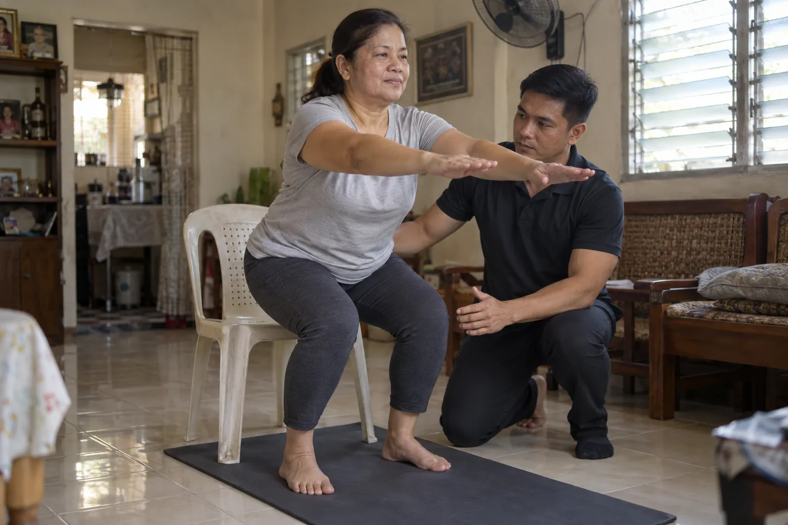 A coach kneels beside a woman in her fifties doing a sit-to-stand squat from a plastic chair in her living room