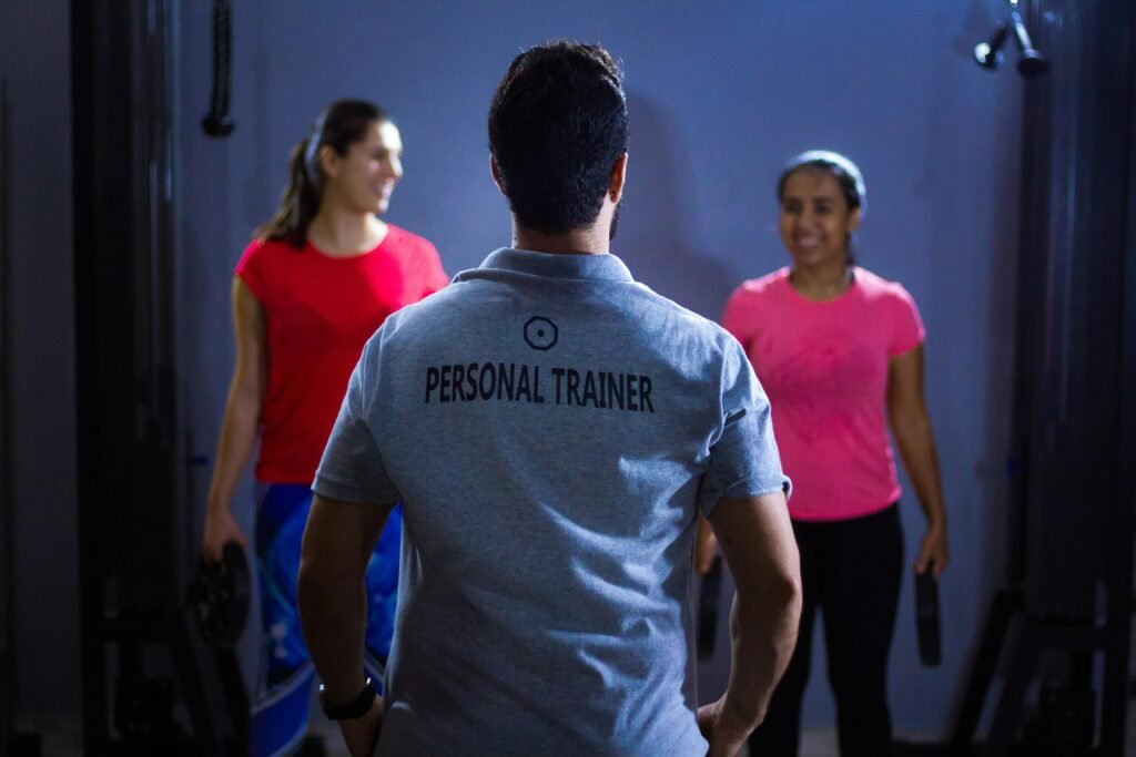 Back view of a personal trainer guiding two women during a gym session indoors.